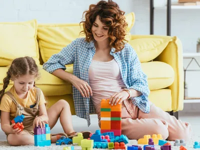 Une nounou souriante et un adorable enfant jouant avec des blocs de construction multicolores sur le sol.
